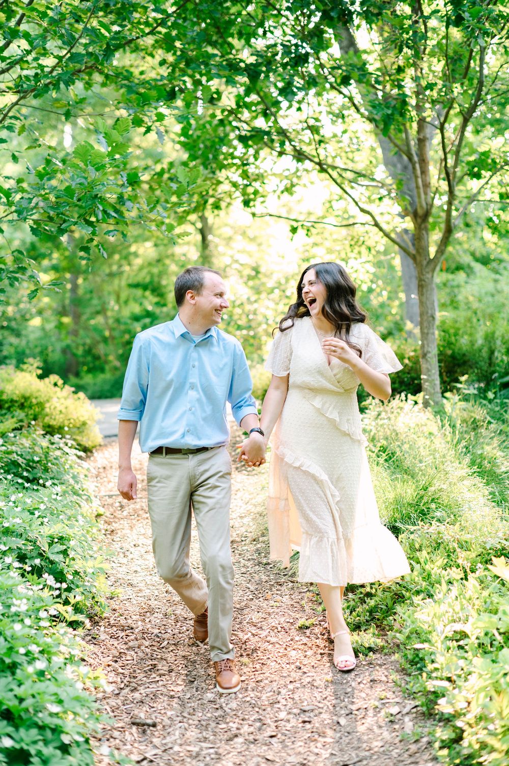 A romantic couple strolling hand in hand along a wooded trail surrounded by green foliage.