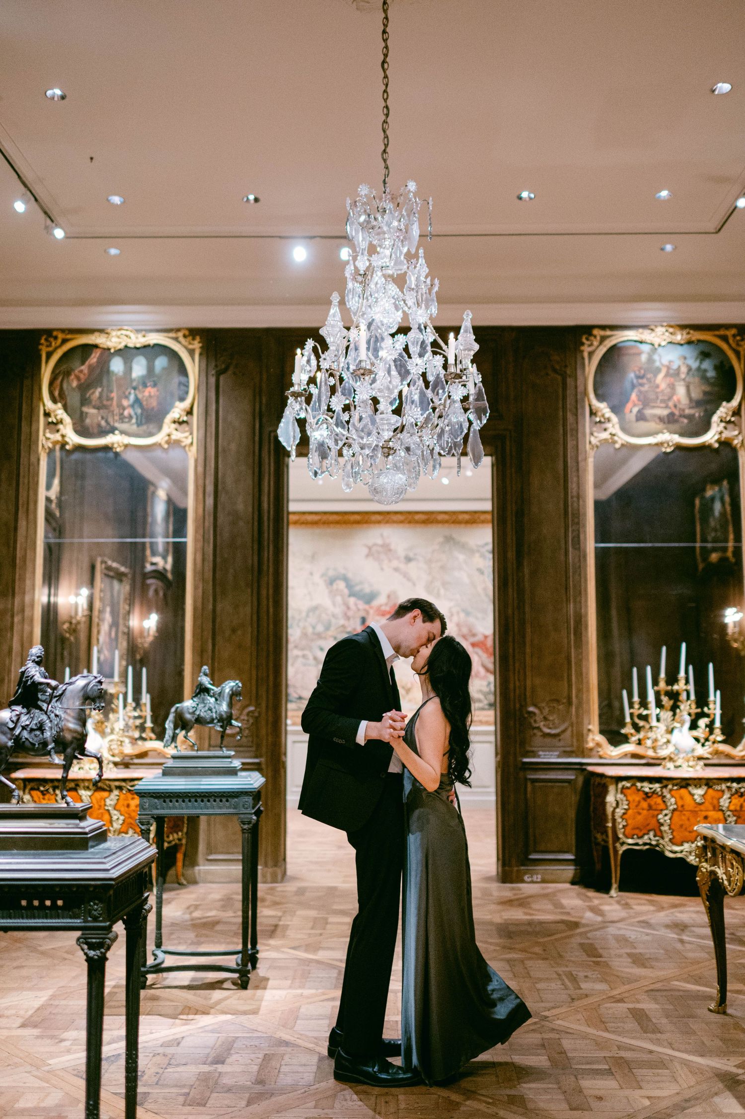 A romantic moment under an elegant crystal chandelier in an ornate ballroom with gilded decor.