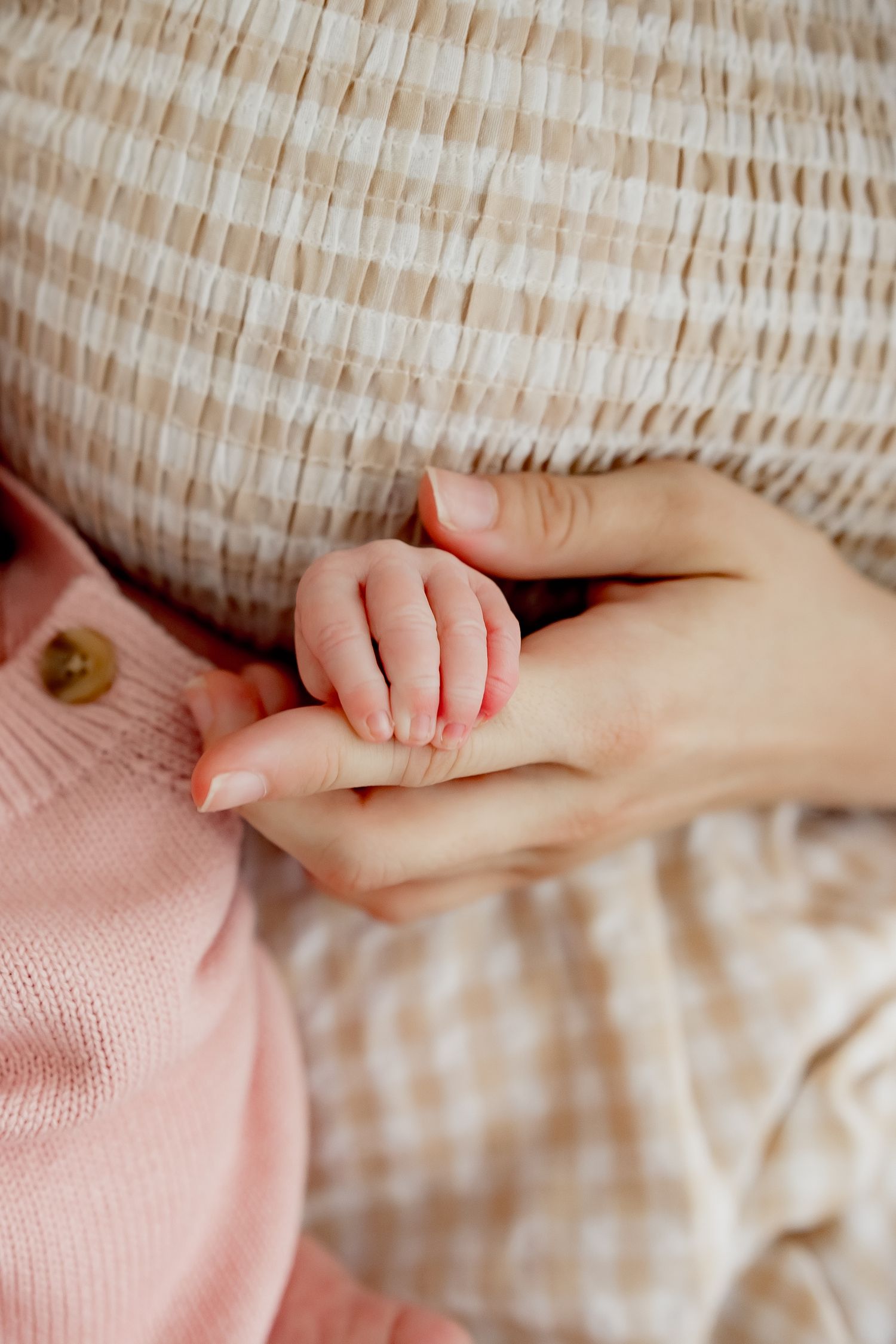 A tiny baby hand gently grasping an adult's finger against a soft pink and beige textured background.