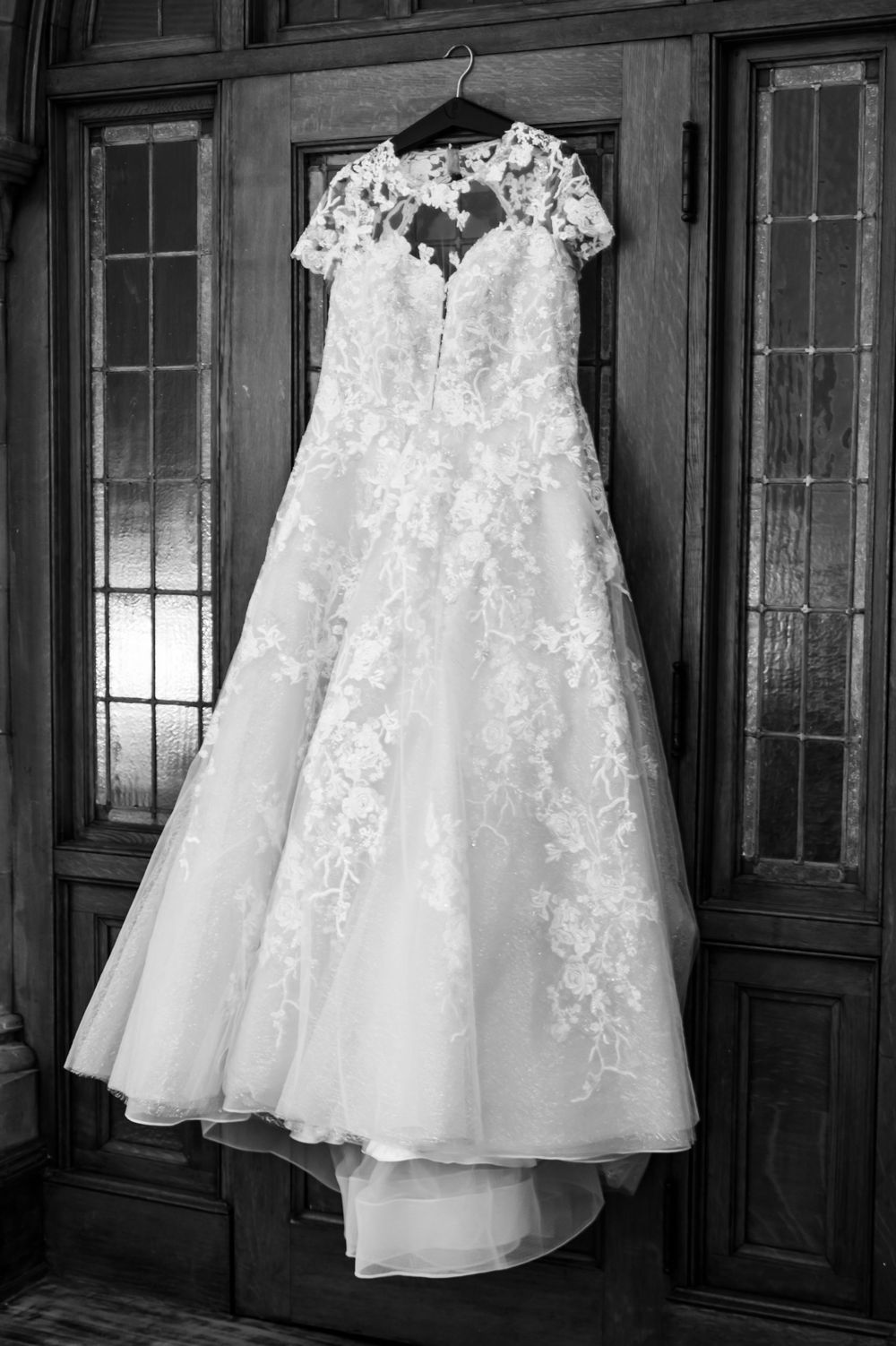 Five delicate white lace wedding dresses with tulle details hang in a row against dark wooden panels in black and white.