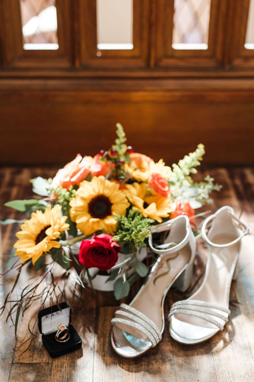 Wedding shoes and floral arrangements with sunflowers and roses displayed on a wooden surface.