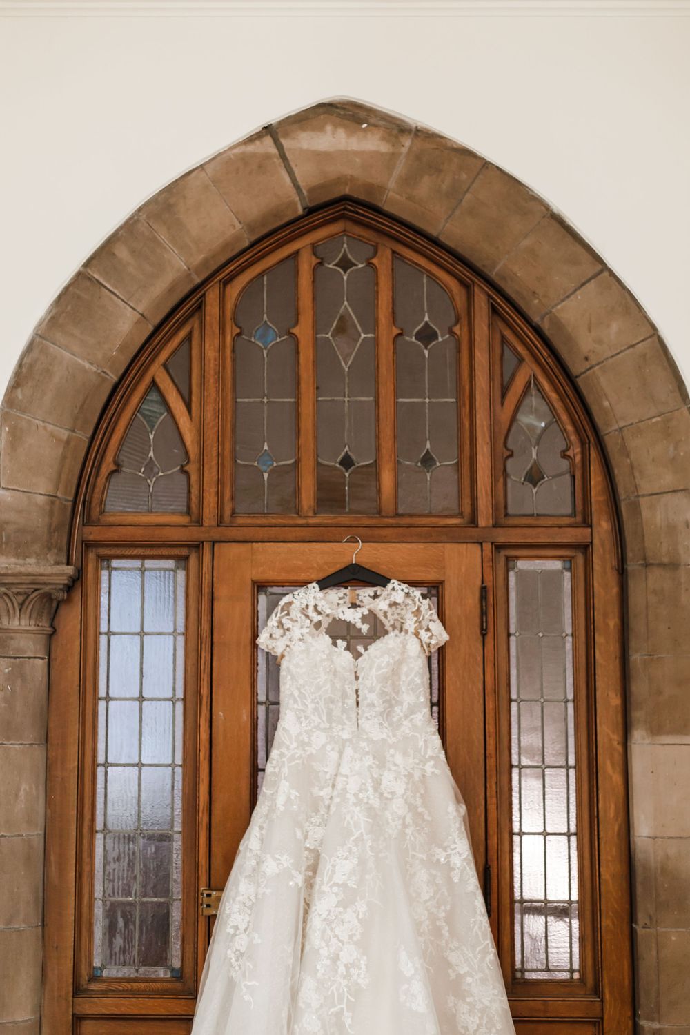 An elegant white lace wedding dress hangs on display against an arched Gothic-style church window with stained glass details.
