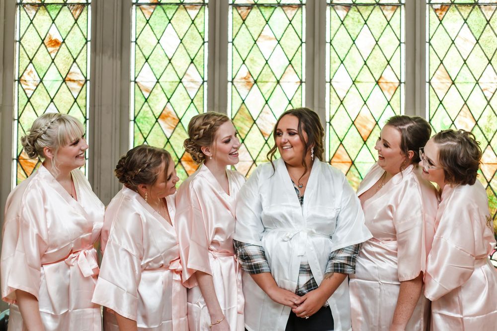 A group in matching light pink satin robes share laughter and conversation while getting ready together.