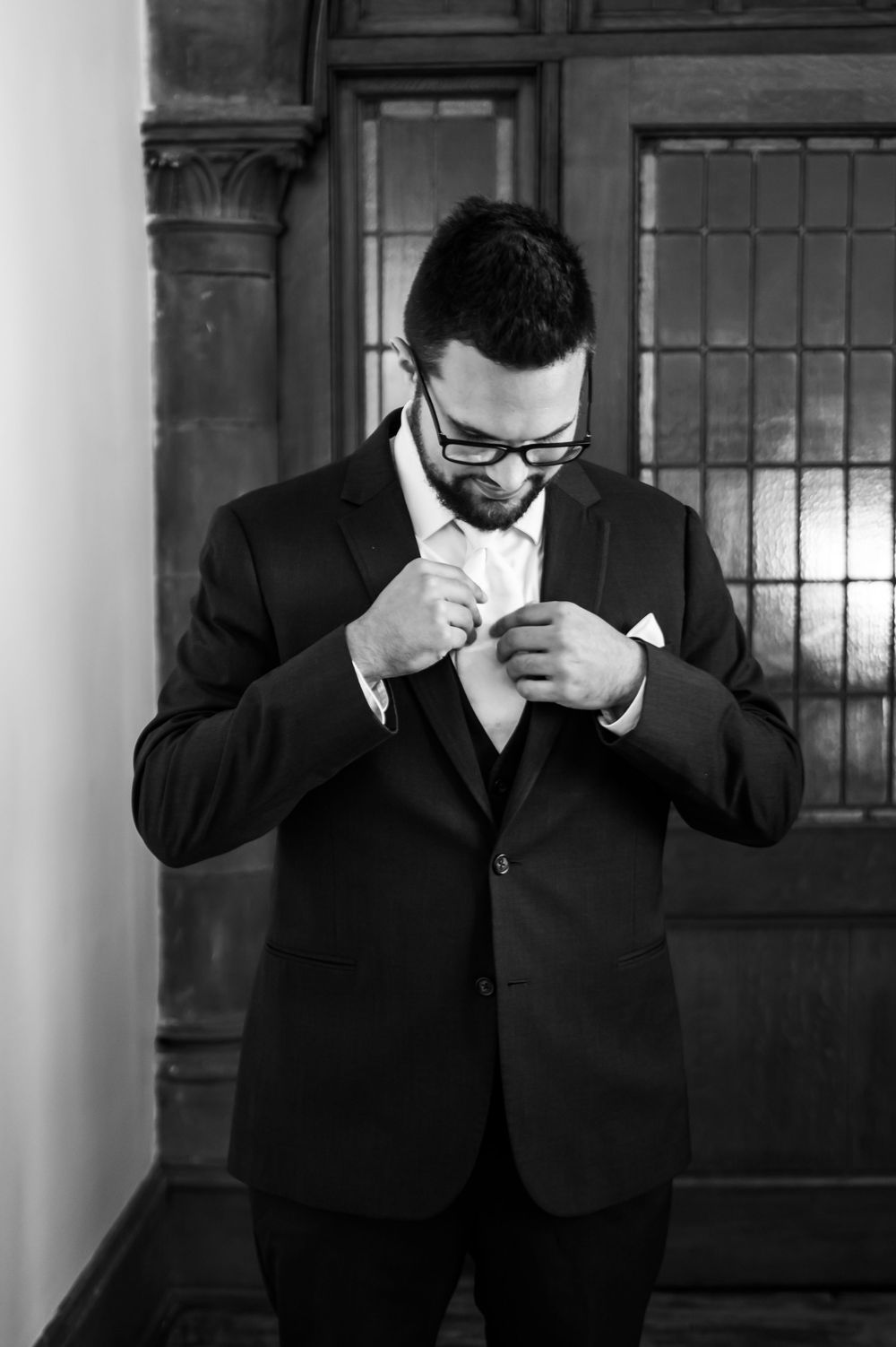 A groom in a black suit and glasses adjusts his tie before the wedding ceremony in a dramatic black and white shot.