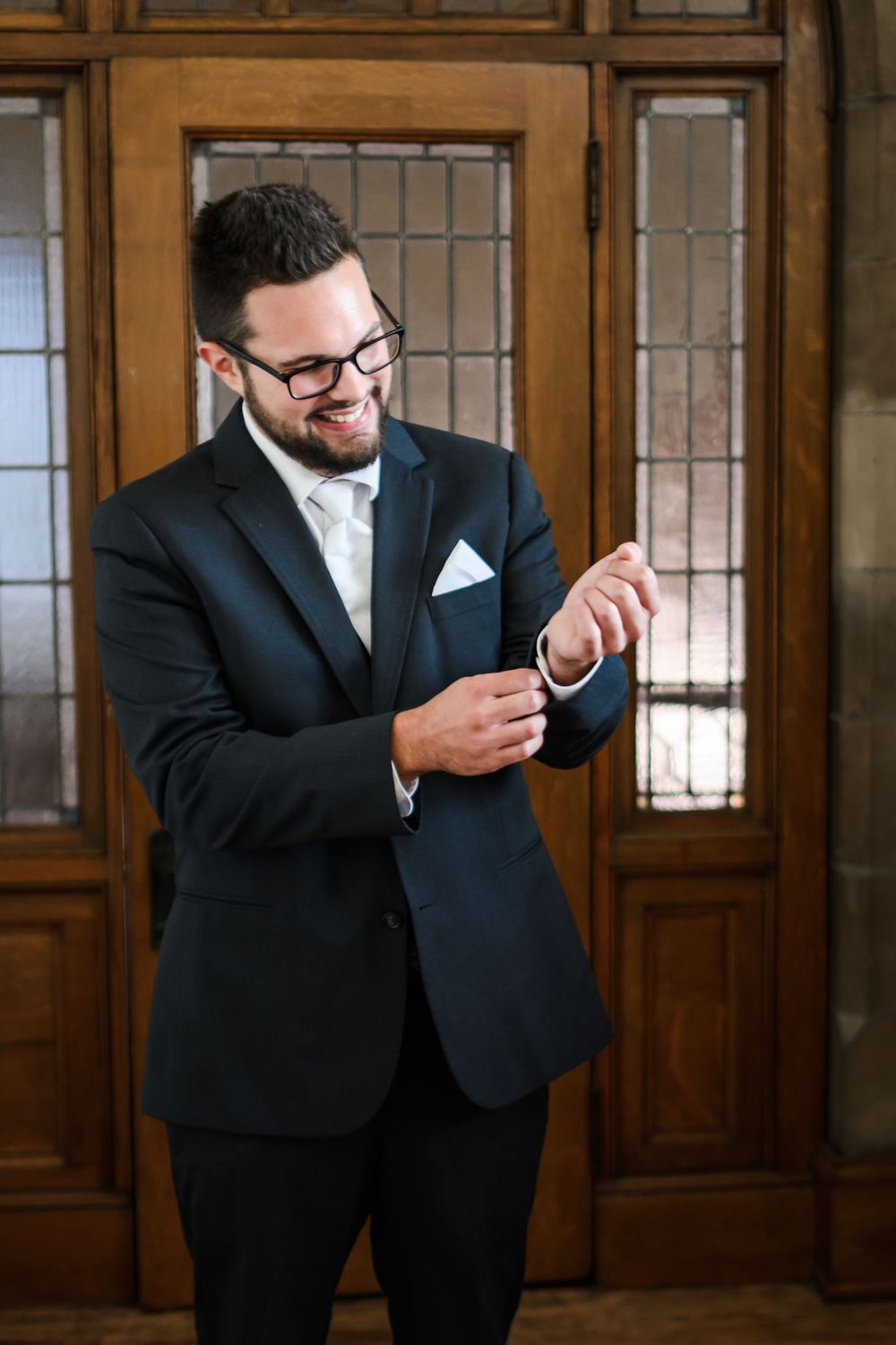 Groom in black suit and glasses checking his phone in an elegant wood-paneled room.