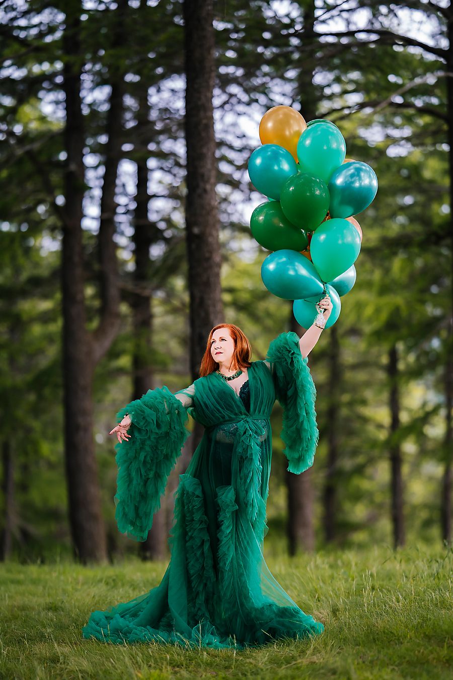 A person wearing an emerald green tulle gown poses with teal balloons in a forest setting.