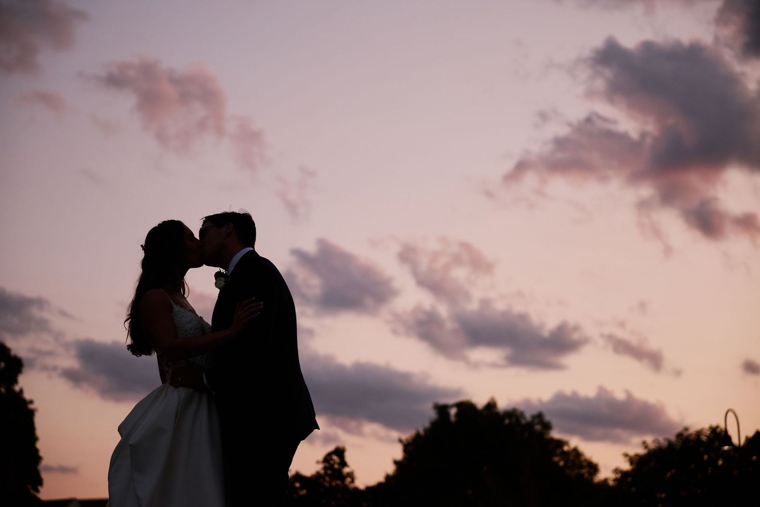 A silhouetted couple shares an intimate moment against a dramatic pink and orange sunset sky in the fall light.