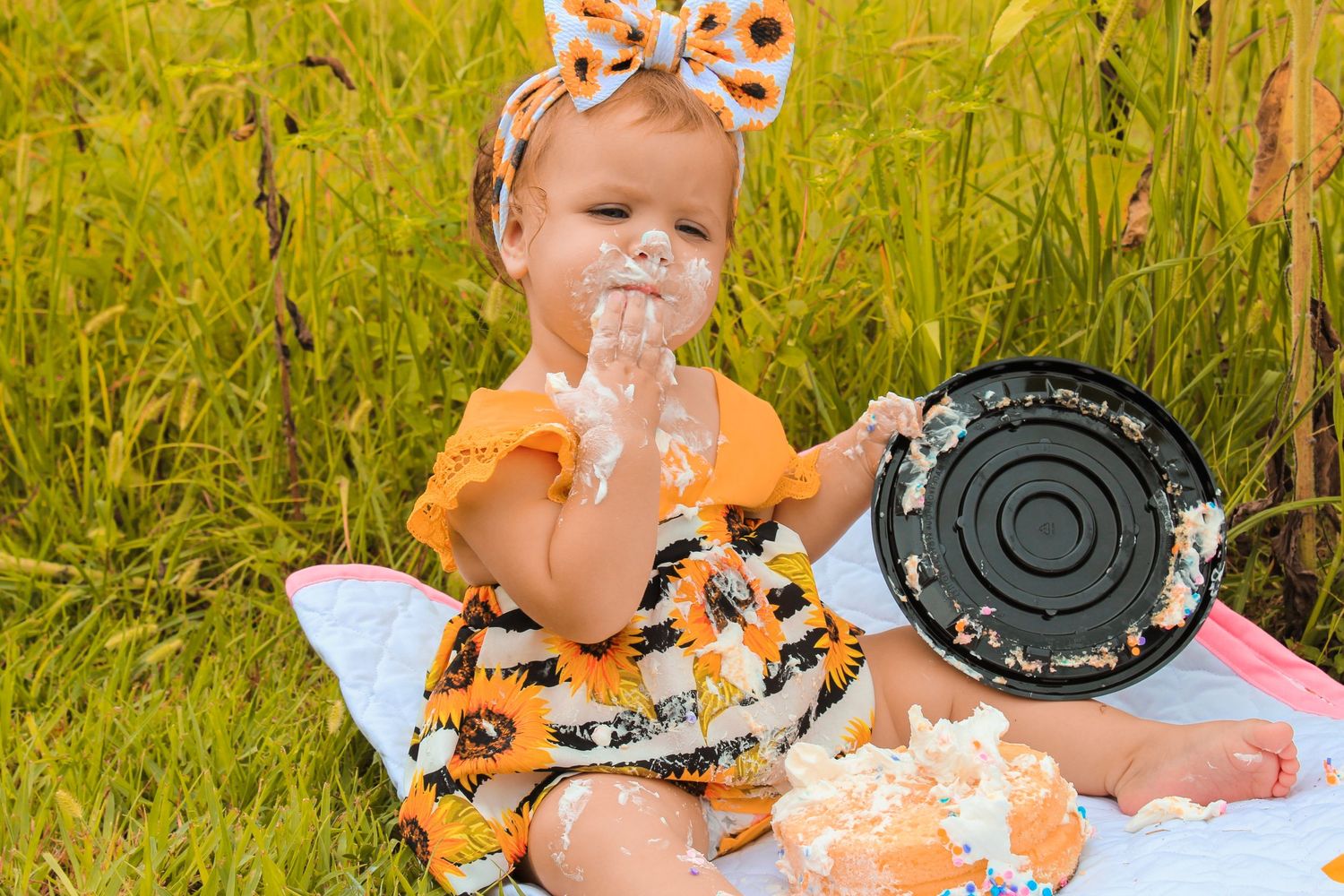 A cake smashing photo session in a golden field shows a baby in orange outfit playing with white frosted cake.