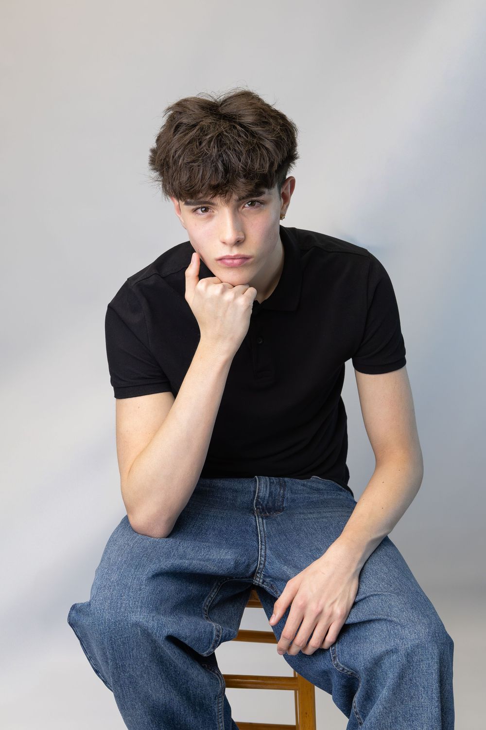 Young person in black t-shirt and jeans sitting thoughtfully on chair against gray background.