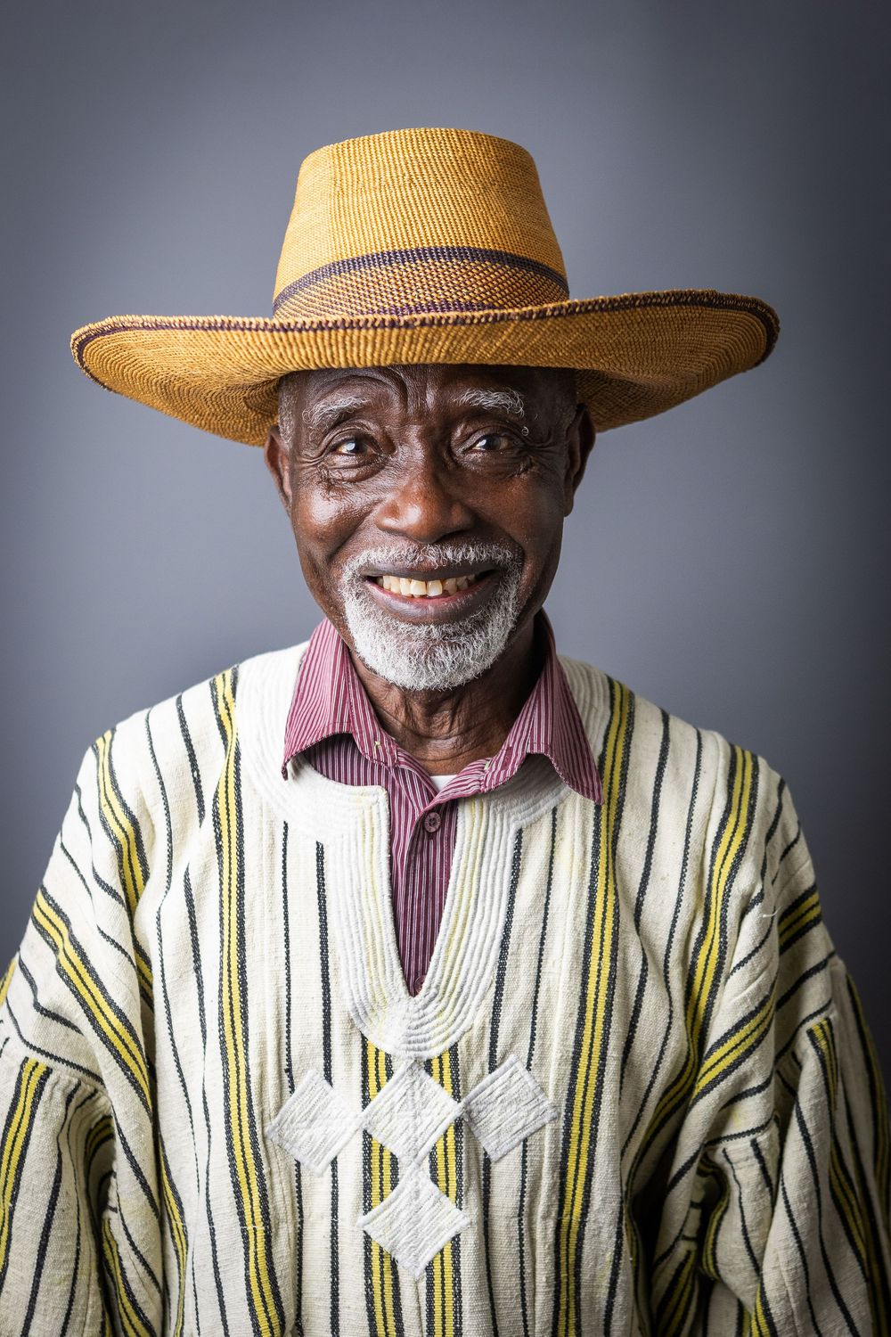 Senior man in striped traditional clothing and straw hat smiling warmly against gray backdrop.