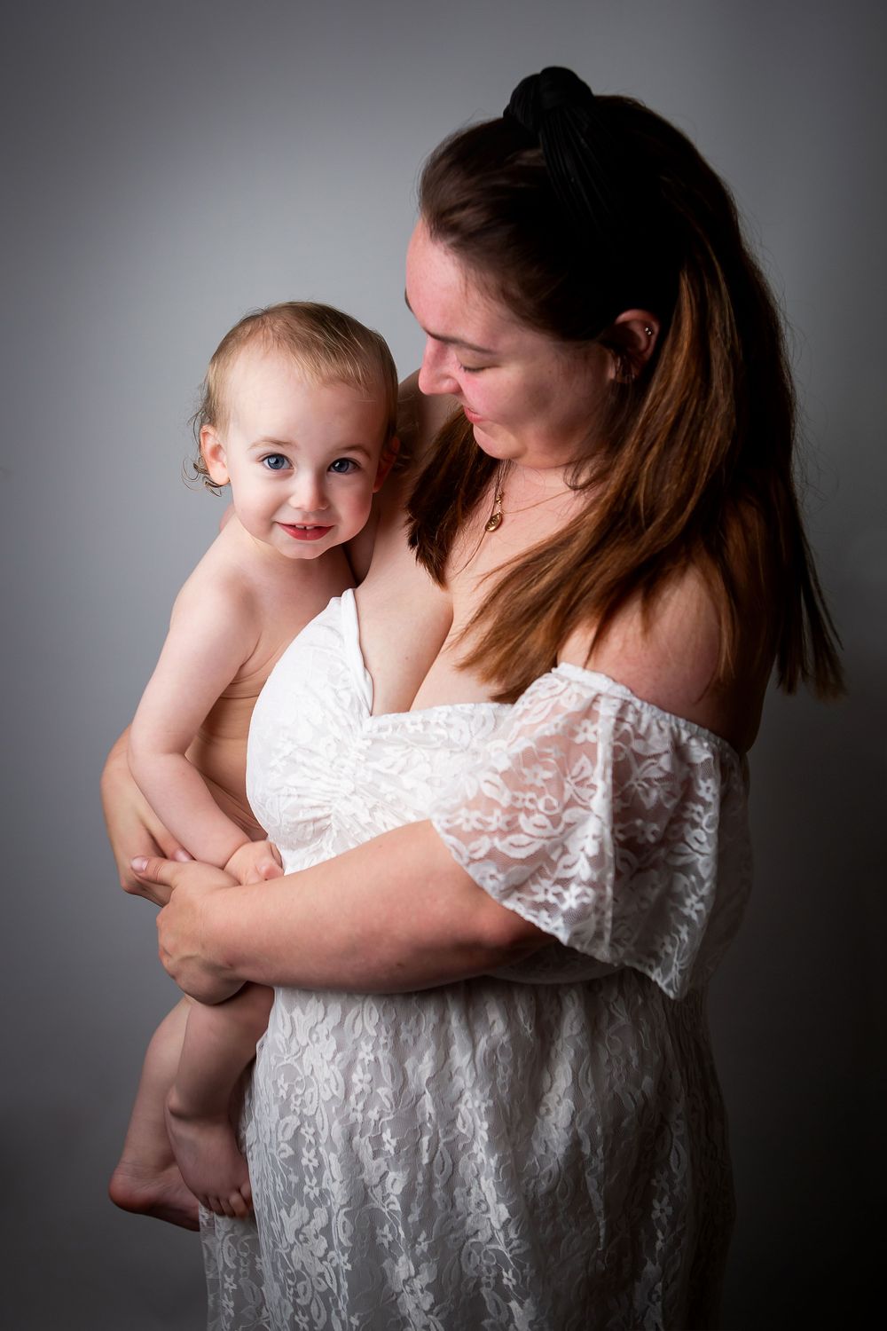 Mother and baby portrait series in white lace dress against dark grey backdrop.