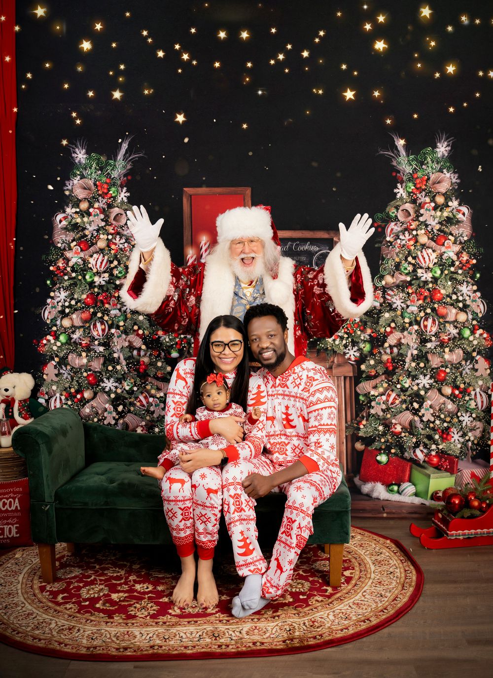 Family in matching red and white patterned holiday pajamas poses against a festive backdrop with Christmas trees and lights.