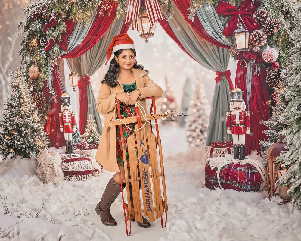 Girl in Santa hat poses with sled in festive Christmas backdrop with nutcrackers and snowy decor.