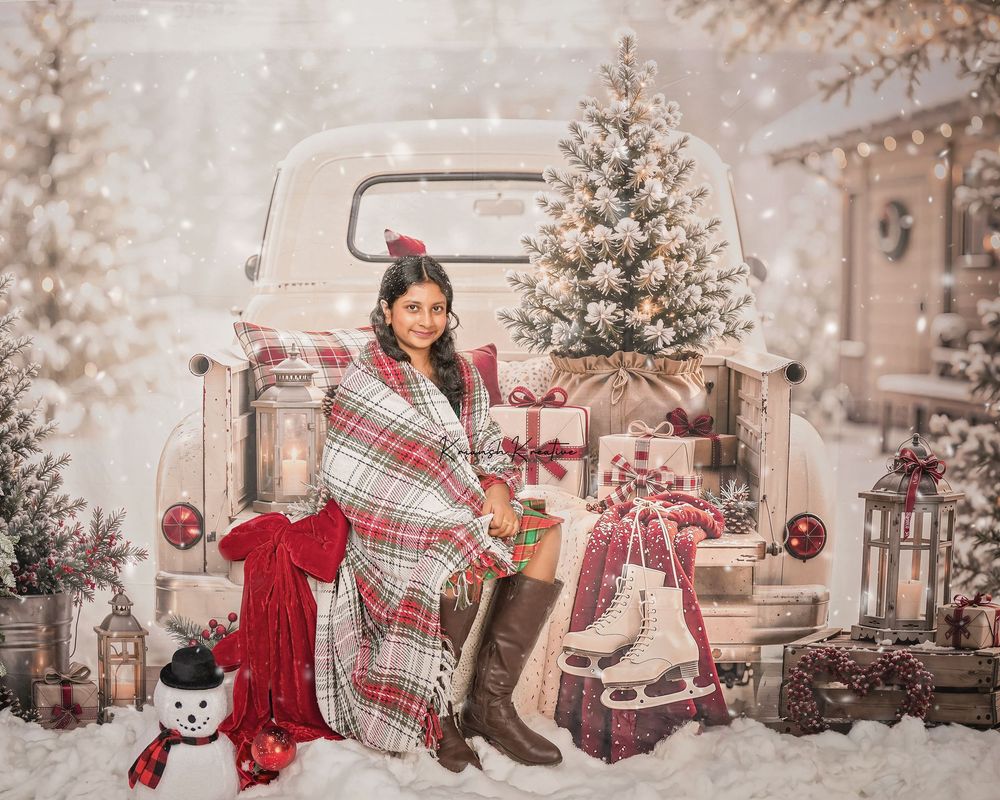 Woman wrapped in plaid blanket sits by vintage white truck with Christmas trees, gifts, and snowman in snowy holiday scene.