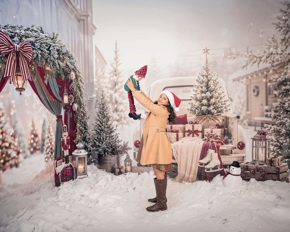 Woman in Santa hat lifts baby in snowy Christmas scene with decorated trees, vintage truck, and holiday gifts.
