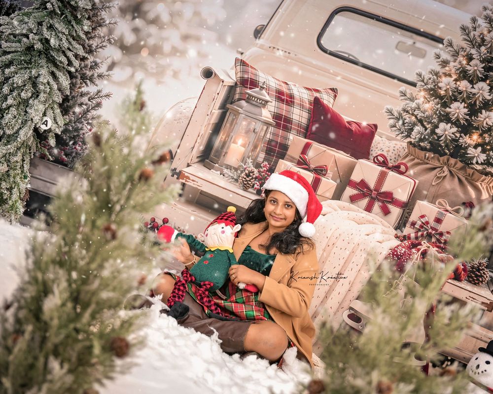 Woman in Santa hat sits with Christmas stuffed toy near snowy trees and vintage truck with holiday gifts.