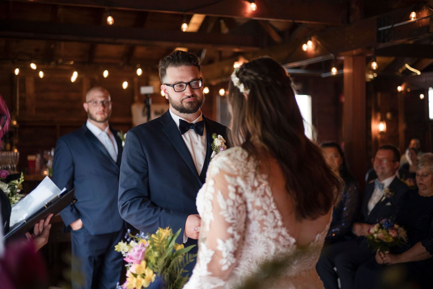An intimate wedding ceremony moment at a rustic indoor venue with warm string lights and wooden beams overhead.