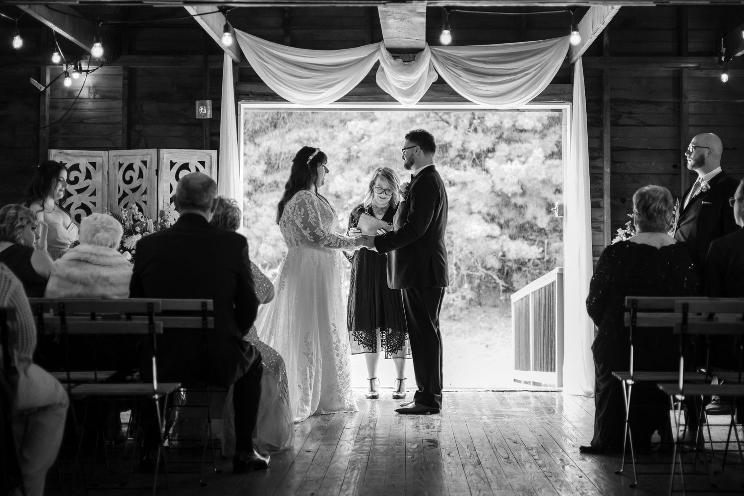 A black and white photograph of a wedding ceremony in a rustic barn venue with draped fabric and string lights.