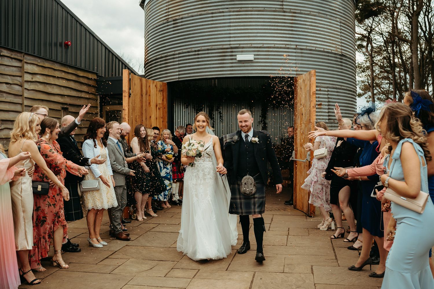 A bride and groom pose by a loch in Scotland, she in a white gown, he in a traditional kilt, with a castle behind.