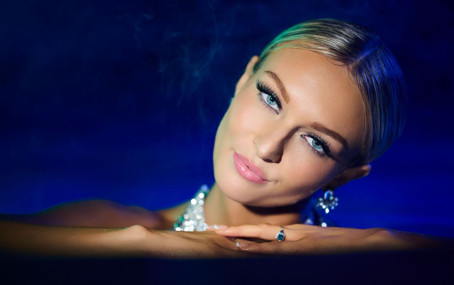 Glamour portrait with dramatic blue lighting shows subject resting on reflective surface wearing sparkly jewelry.