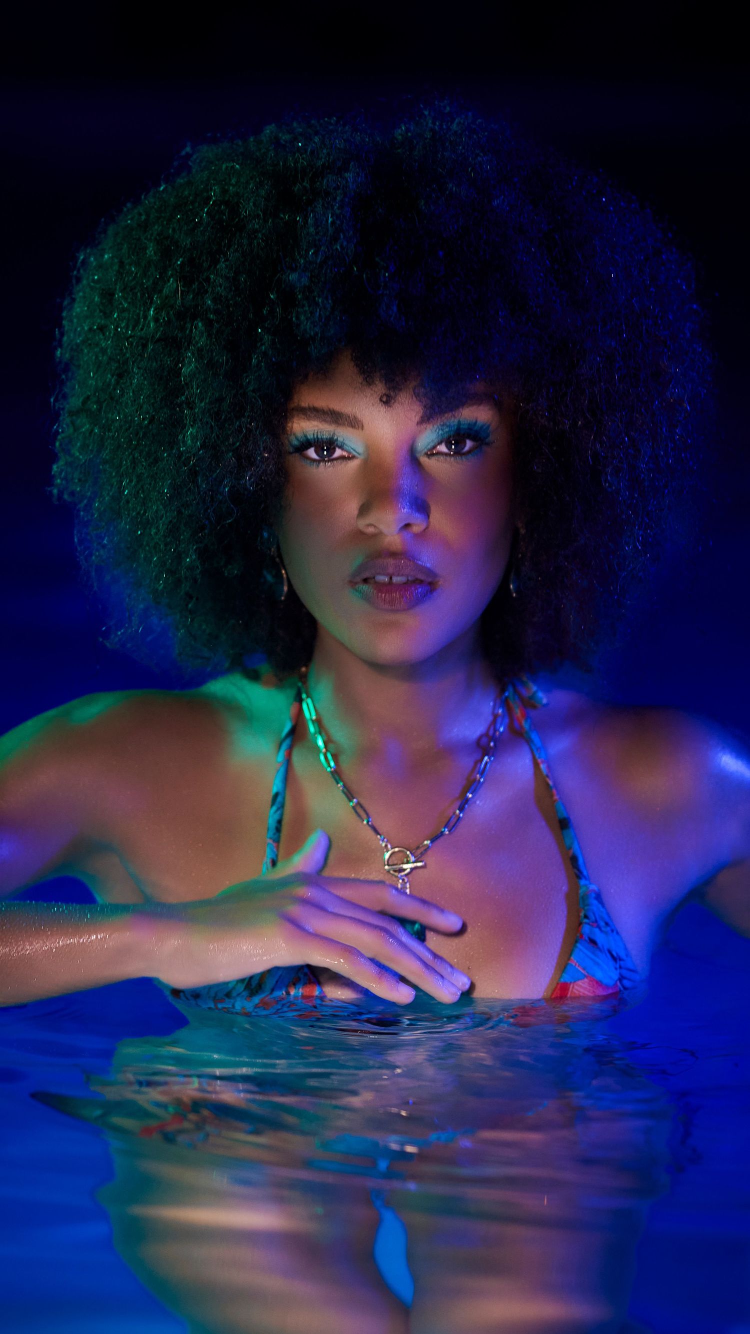 A model poses in a dramatic underwater photoshoot with neon blue lighting and natural curly hair.