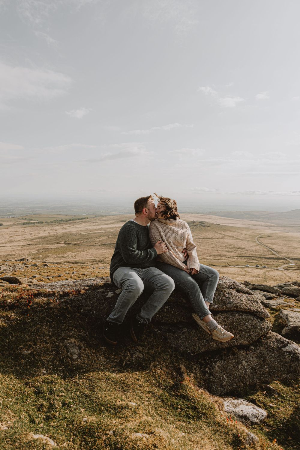 A couple sits together on a rocky outcrop overlooking rolling hills and distant mountains.