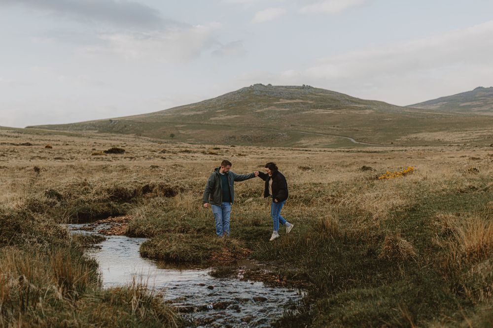 Two people holding hands while crossing a small stream in rugged moorland landscape.