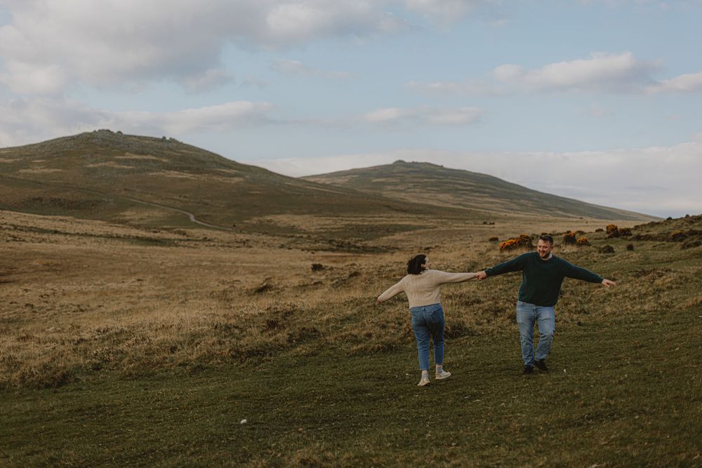 Two people running freely through green hills under cloudy skies.