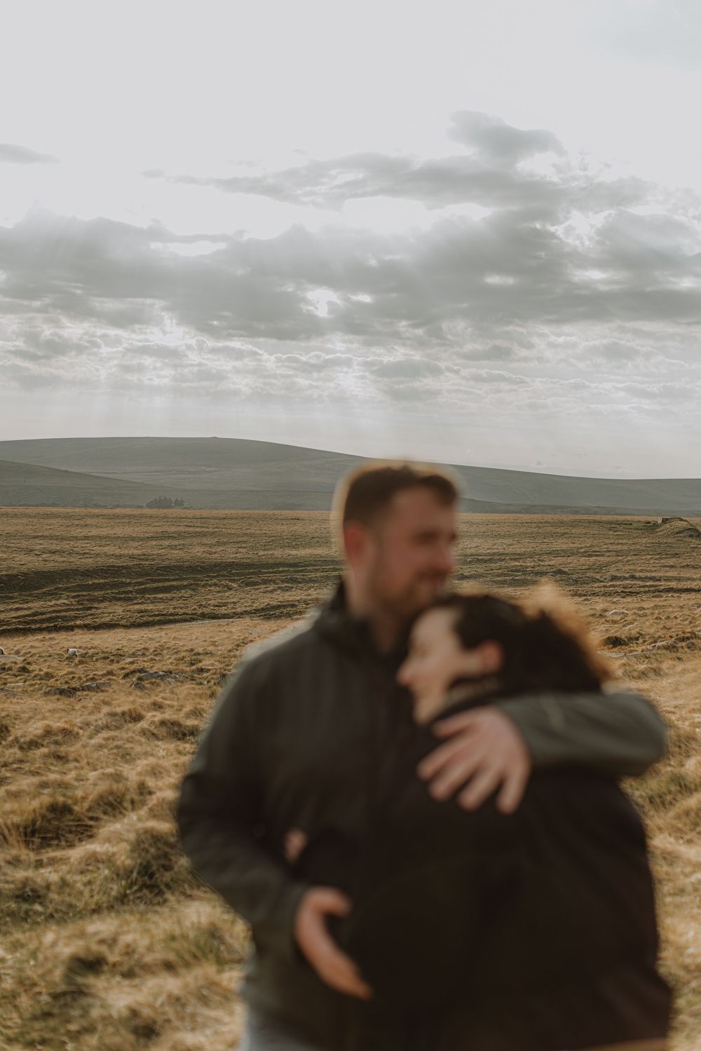 A couple embraces in a tender moment against a backdrop of rolling hills and moody skies.