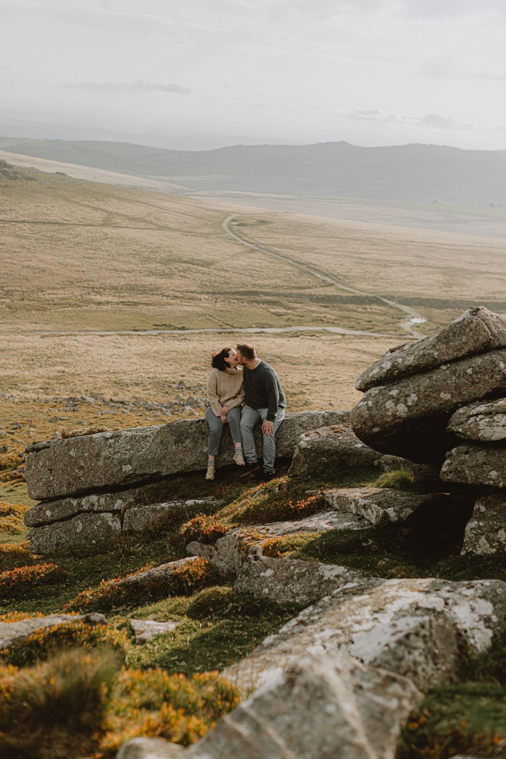 A couple shares an intimate moment while sitting on rocky outcrops overlooking a vast valley.