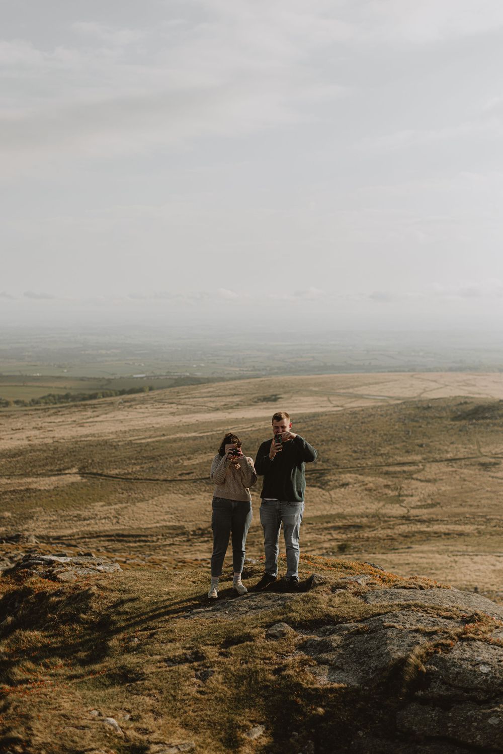 Two people stand on a rocky outcrop overlooking a misty moorland landscape.