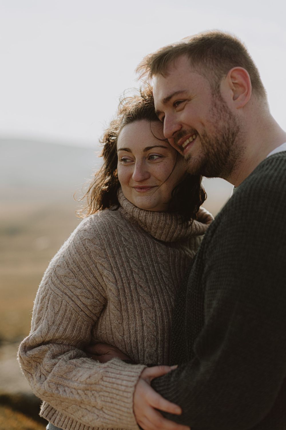 A couple shares a warm embrace while wearing cozy sweaters against a windswept hillside backdrop.