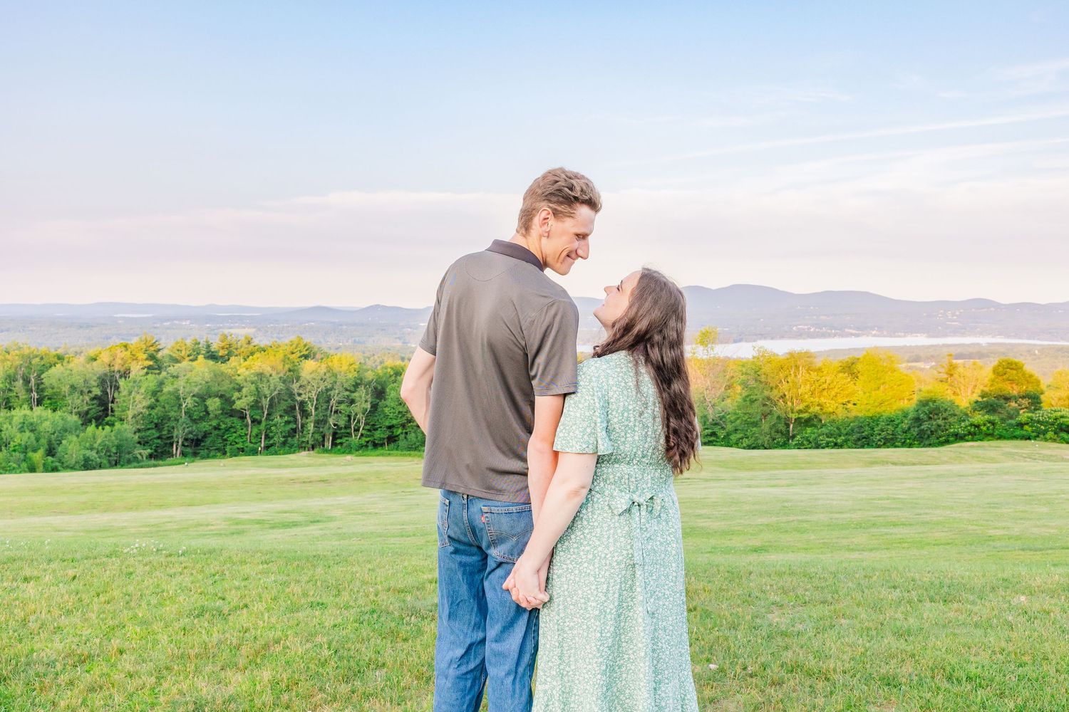 A couple embraces in a scenic field with rolling green hills and golden flowers in the background.