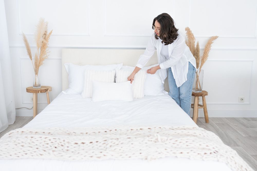 Person making a white bed with decorative throw pillows and pampas grass decor in a bright minimalist bedroom setting.