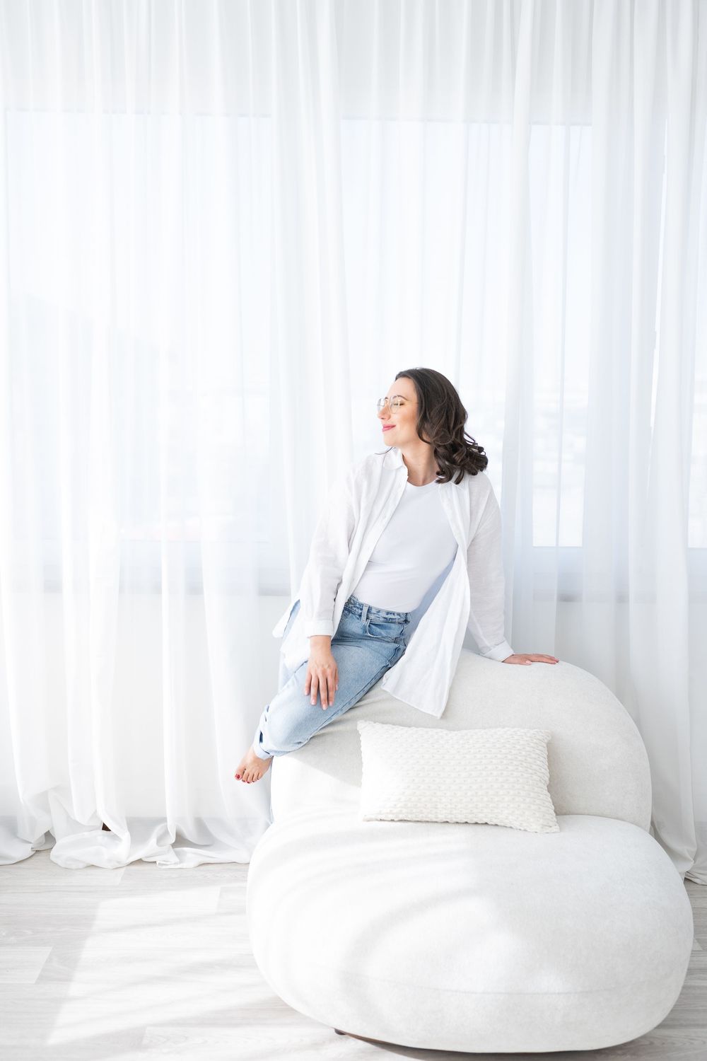 Woman relaxing on a plush white bean bag chair against sheer curtains in a bright contemporary room.