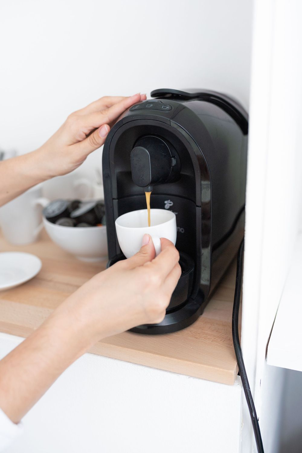Hands operating a black coffee maker machine brewing espresso into white cup.