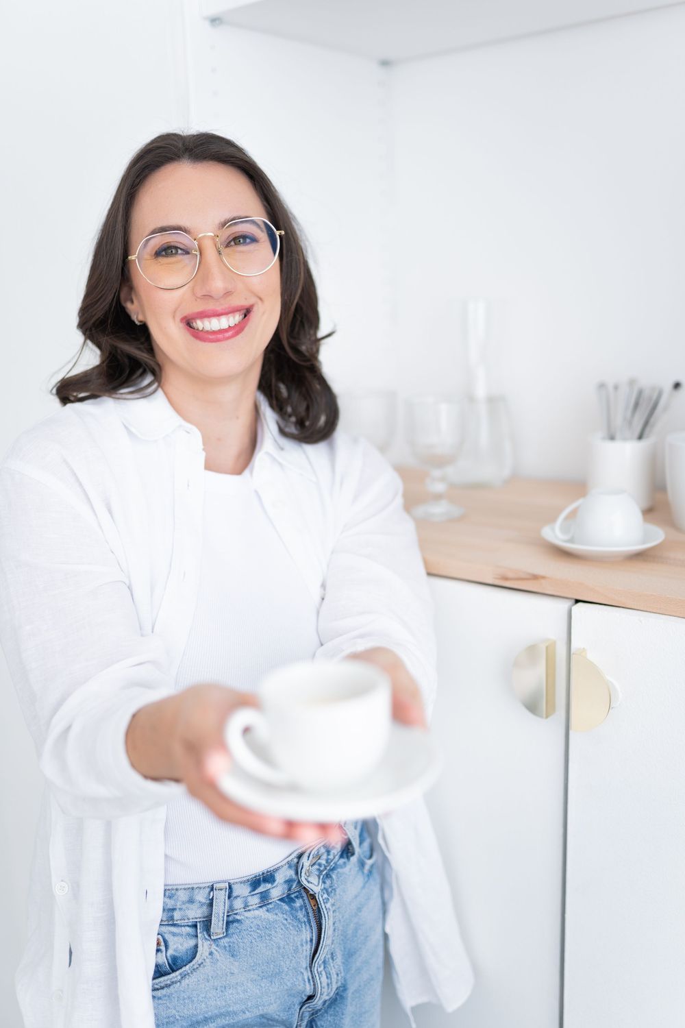 Professional in glasses serving coffee in modern minimalist white kitchen with warm natural lighting.