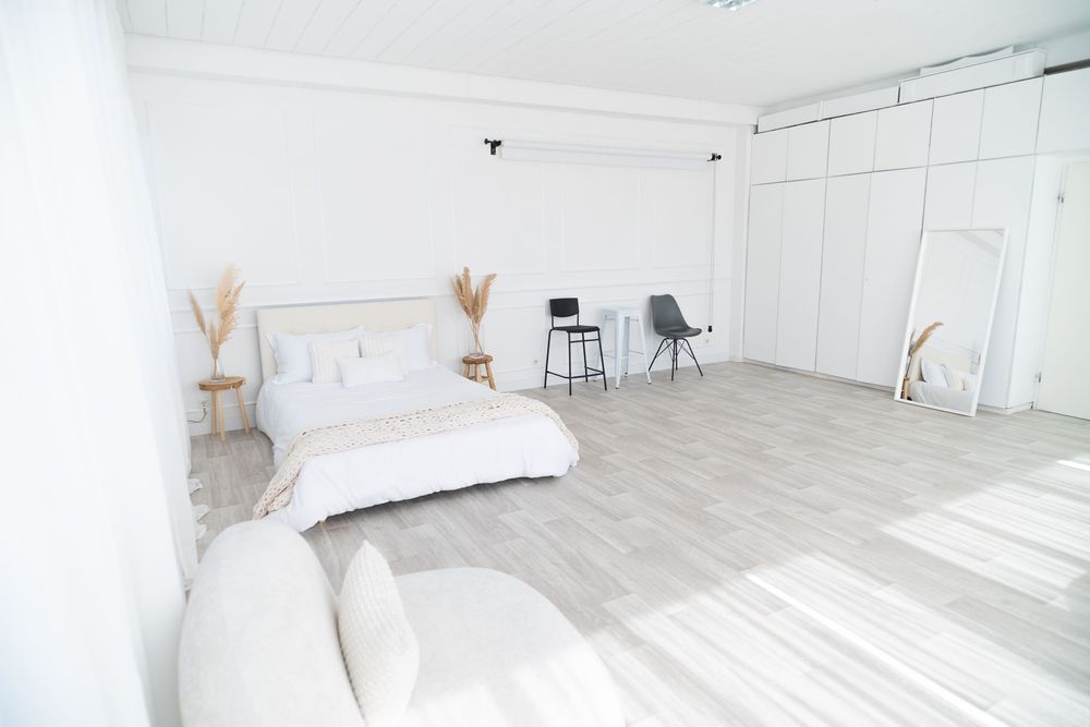 Minimalist white photo studio space with bed setup, white furniture and natural light streaming through windows.