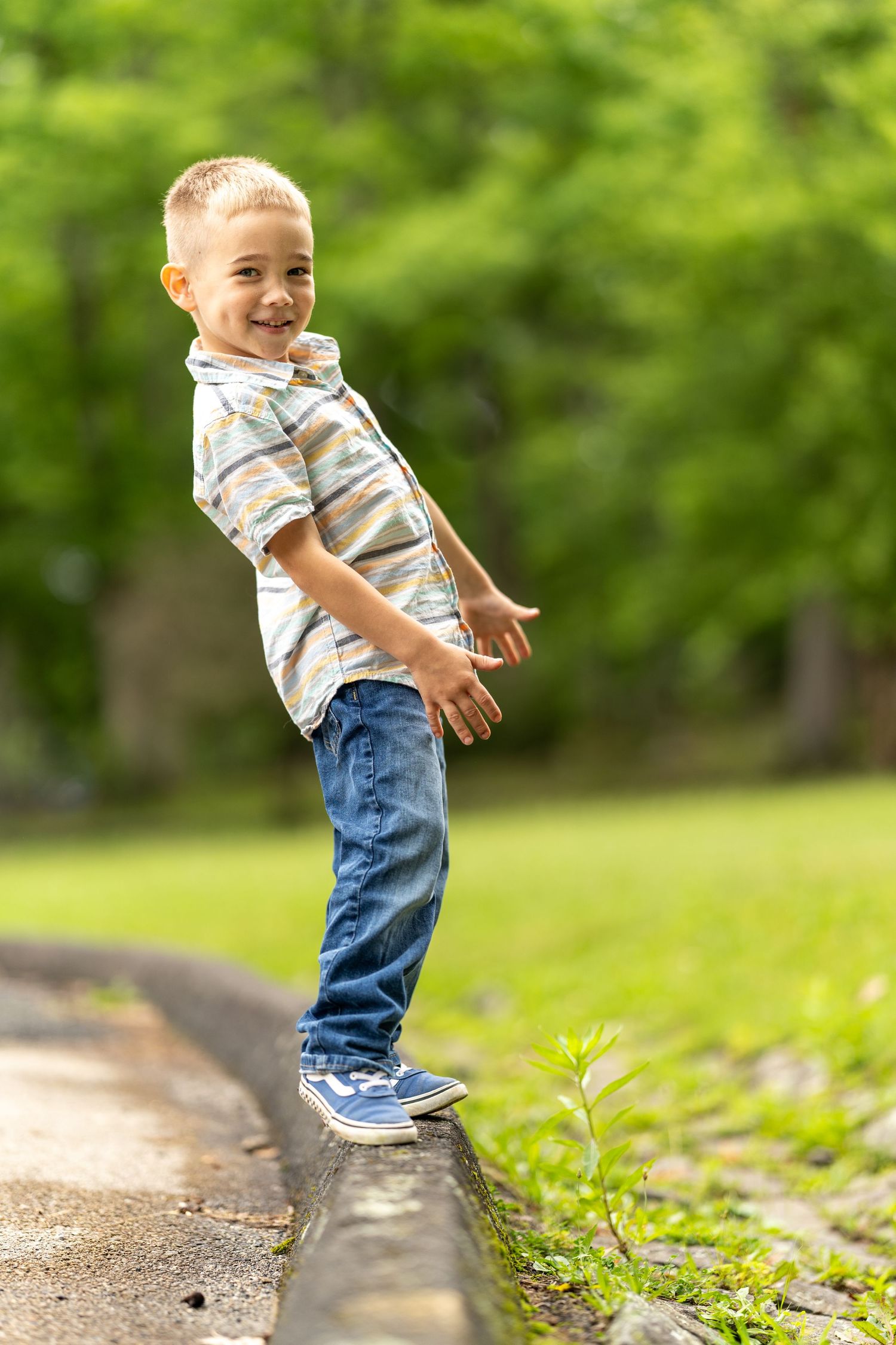 Young child balancing and playing on a concrete curb in a park setting wearing jeans and striped shirt on sunny day.
