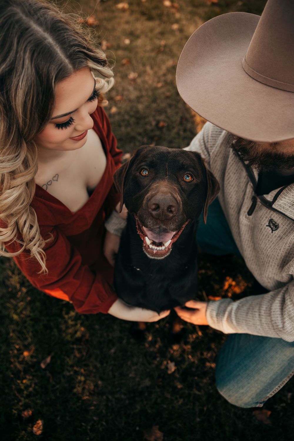Happy black dog poses between couple in red dress and cowboy hat during autumn photoshoot.
