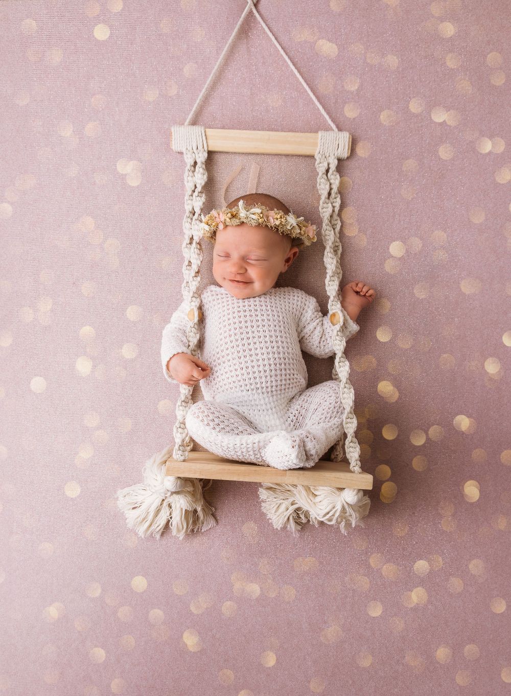 Sleeping newborn baby resting in miniature wooden swing against pink polka dot background.