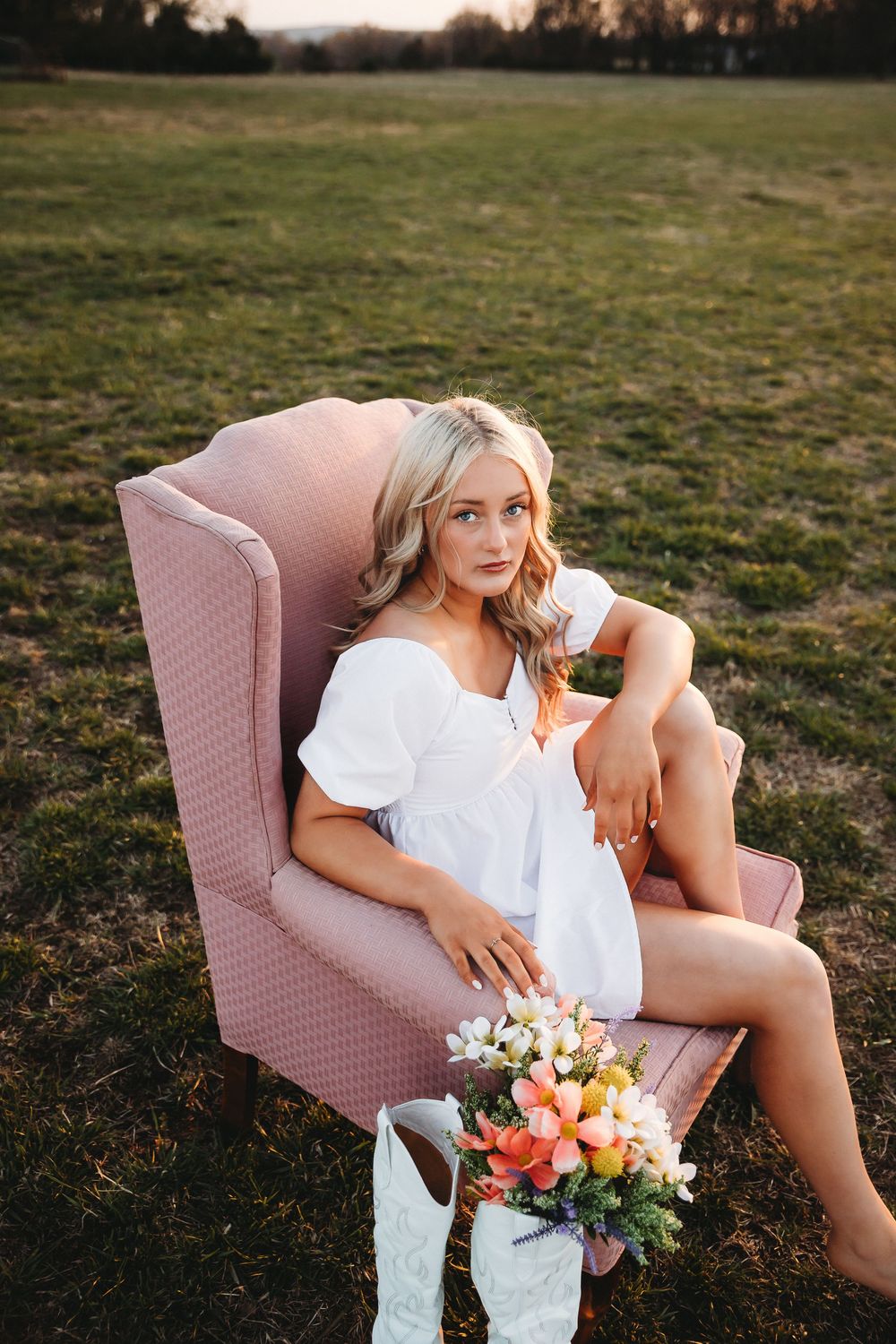 Person in white dress lounging on pink chair with flowers at sunset.