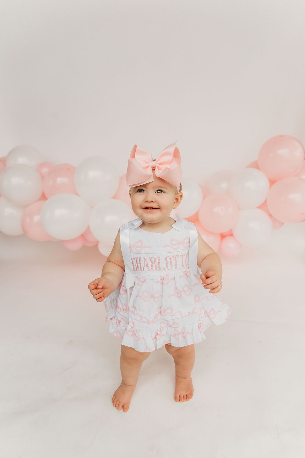 Baby dressed in pink and white outfit stands against pink and white balloon backdrop.