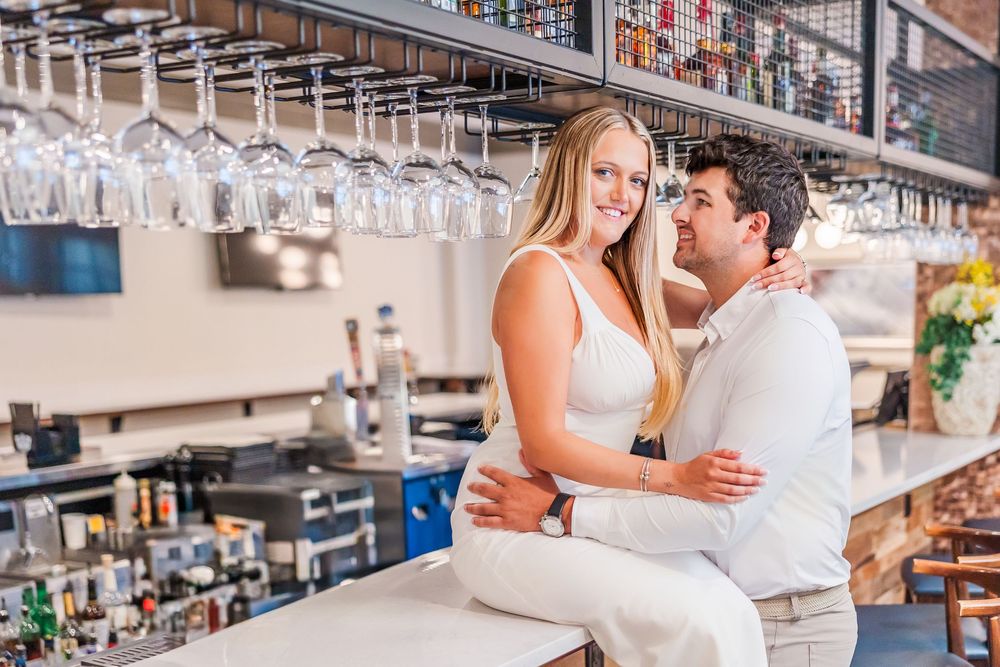 A couple embraces at a bar with hanging wine glasses visible above.