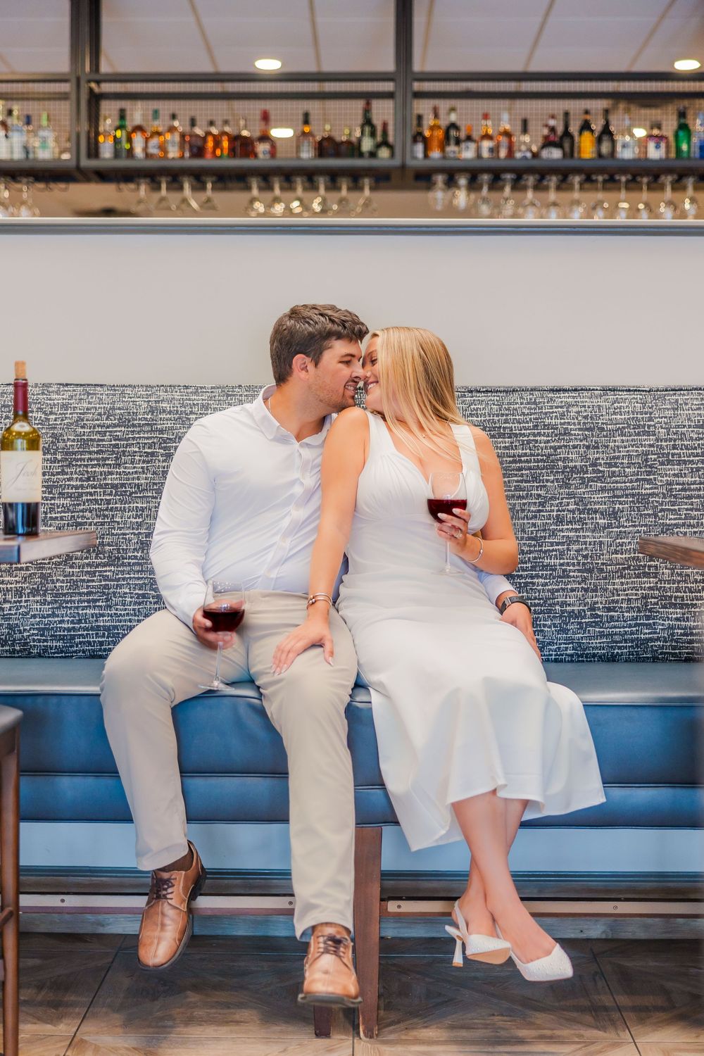 Couple in white outfits share romantic moments while seated on a blue banquette at an upscale bar.