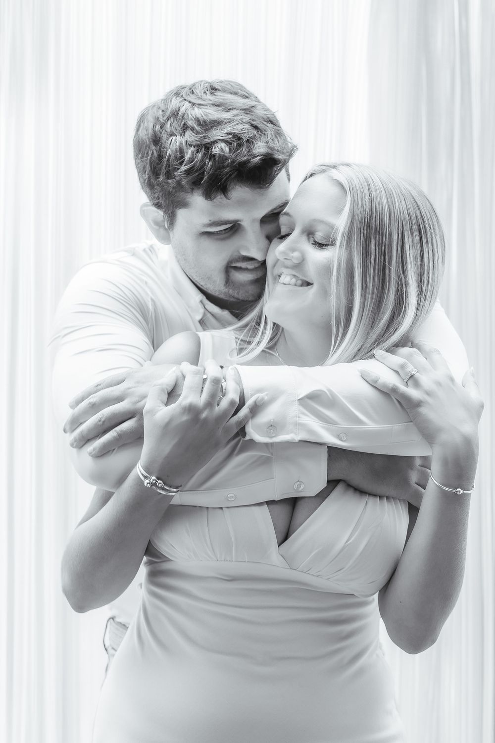 A couple embraces intimately while sharing a joyful moment together against white curtains in black and white.