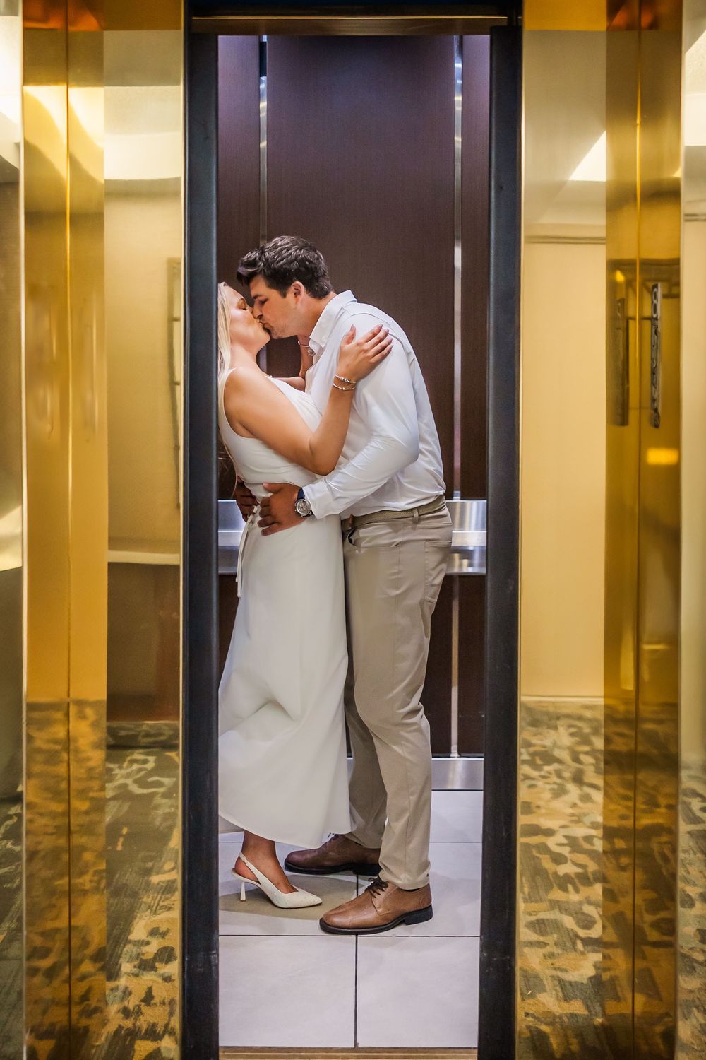 A romantic couple shares an intimate kiss while standing in a modern elevator with gold-toned walls.