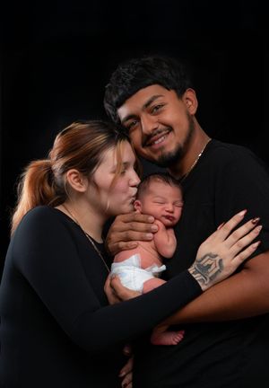 Smiling young parents in black outfits hold their newborn baby as the mother gives a gentle kiss on its cheek.