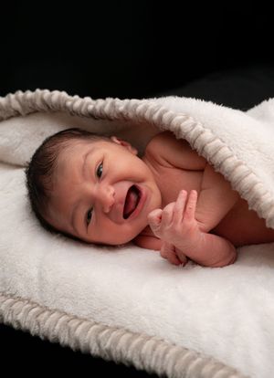 Happy newborn baby smiling and laughing while lying on a fluffy white blanket against a dark background.
