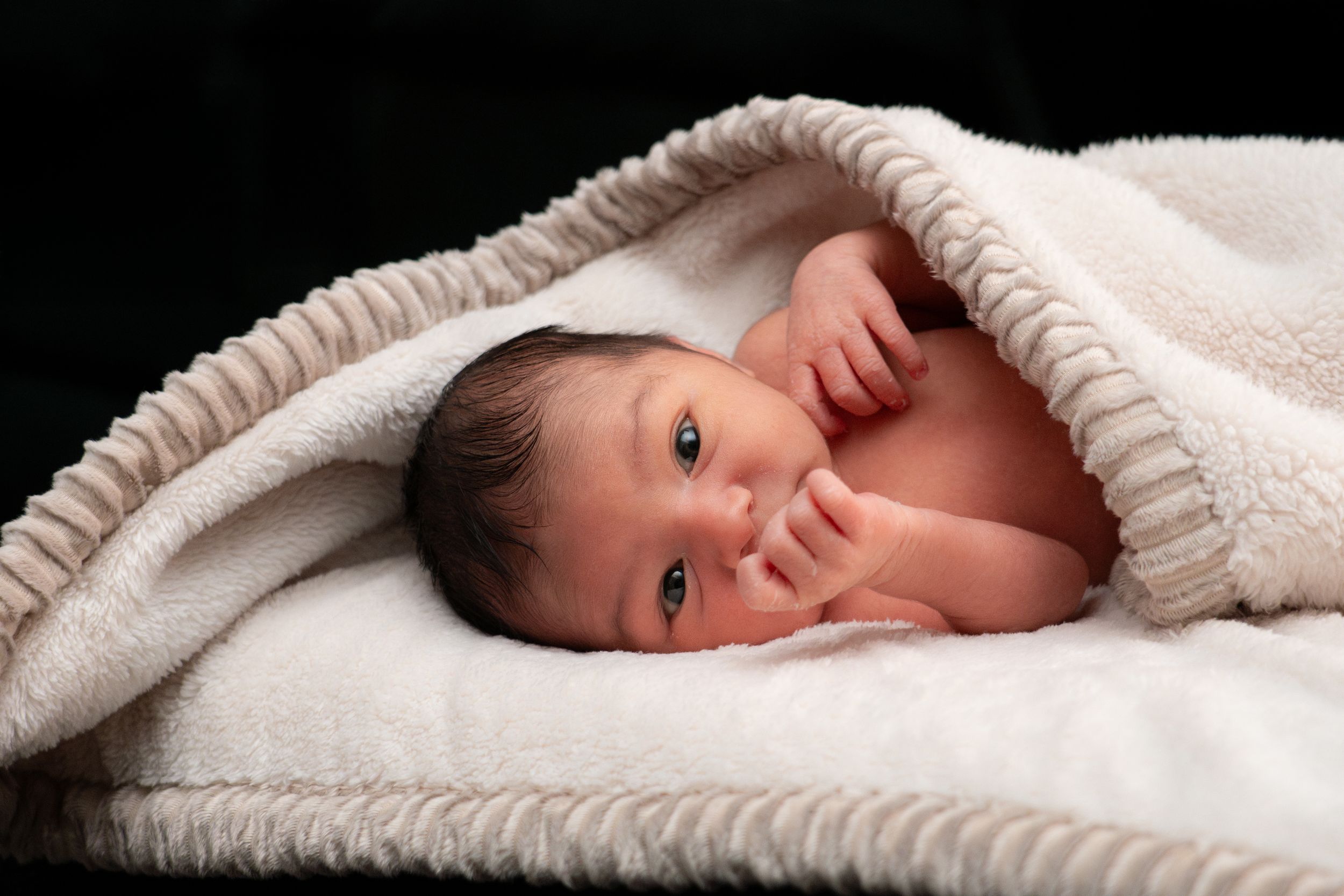 Newborn baby wrapped in a soft white blanket with fingers near mouth on dark background.